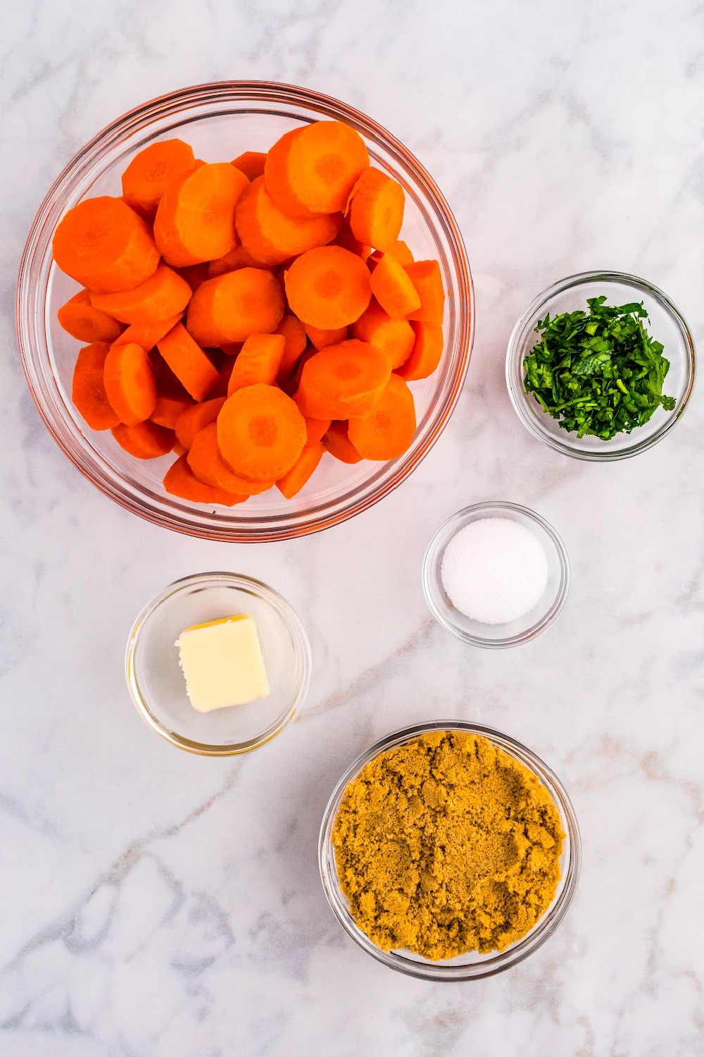 Measured ingredients for making Brown Sugar Carrots presented in clear glass bowls on a marble countertop.