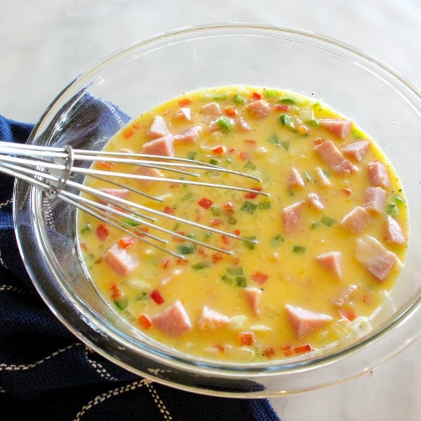 Ham, bell peppers, onions and cheese added to egg mixture in clear glass bowl