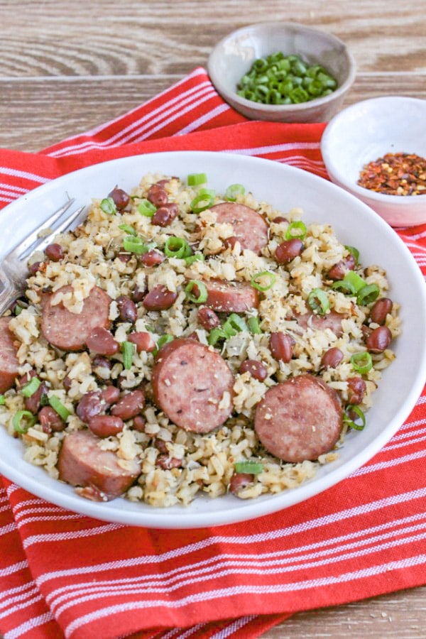 red beans and rice in a bowl with fork with pinch bowls of chopped scallions and red pepper flakes on top of red and white towel with woodent back ground 