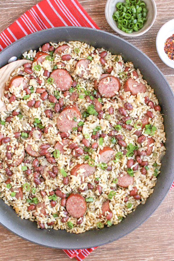 red beans and rice in skillet on top of red and white striped towel with chopped scallions and red pepper flakes in pinch bowls on the right 