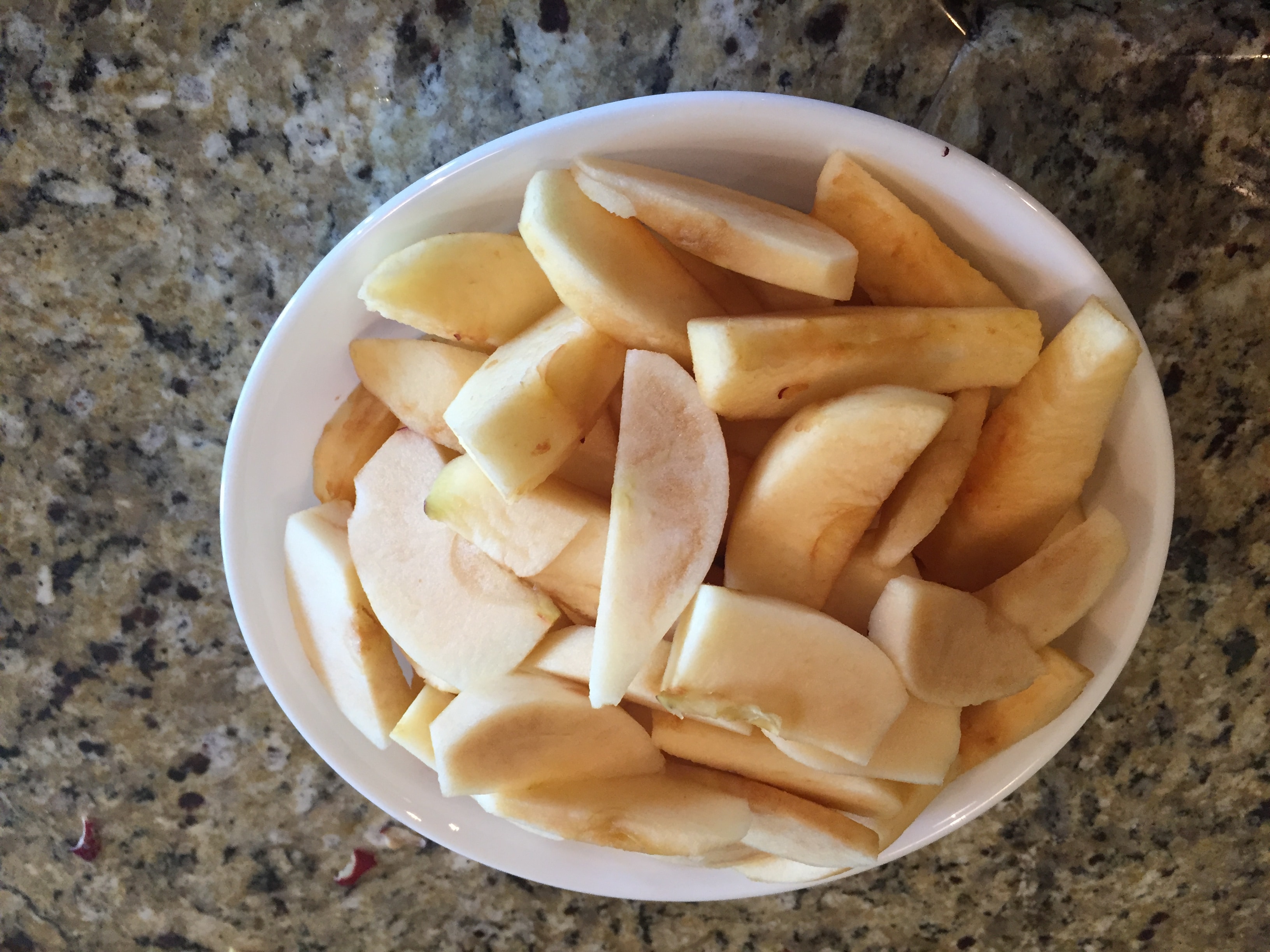 apple slices that have been peeled in a white bowl on tan granite counter top 
