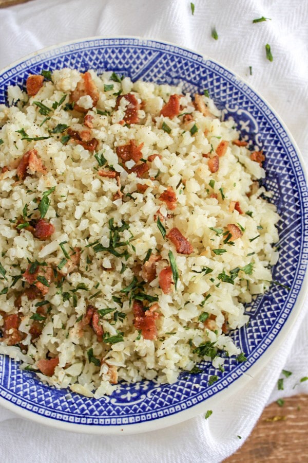  cauliflower rice with bacon and chives in a blue and white bowl sitting on s white dish towel