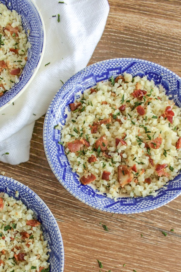 Three blue and white bowls of cauliflower rice with bacon and chive on wooden background with white dish towel under one bowl 