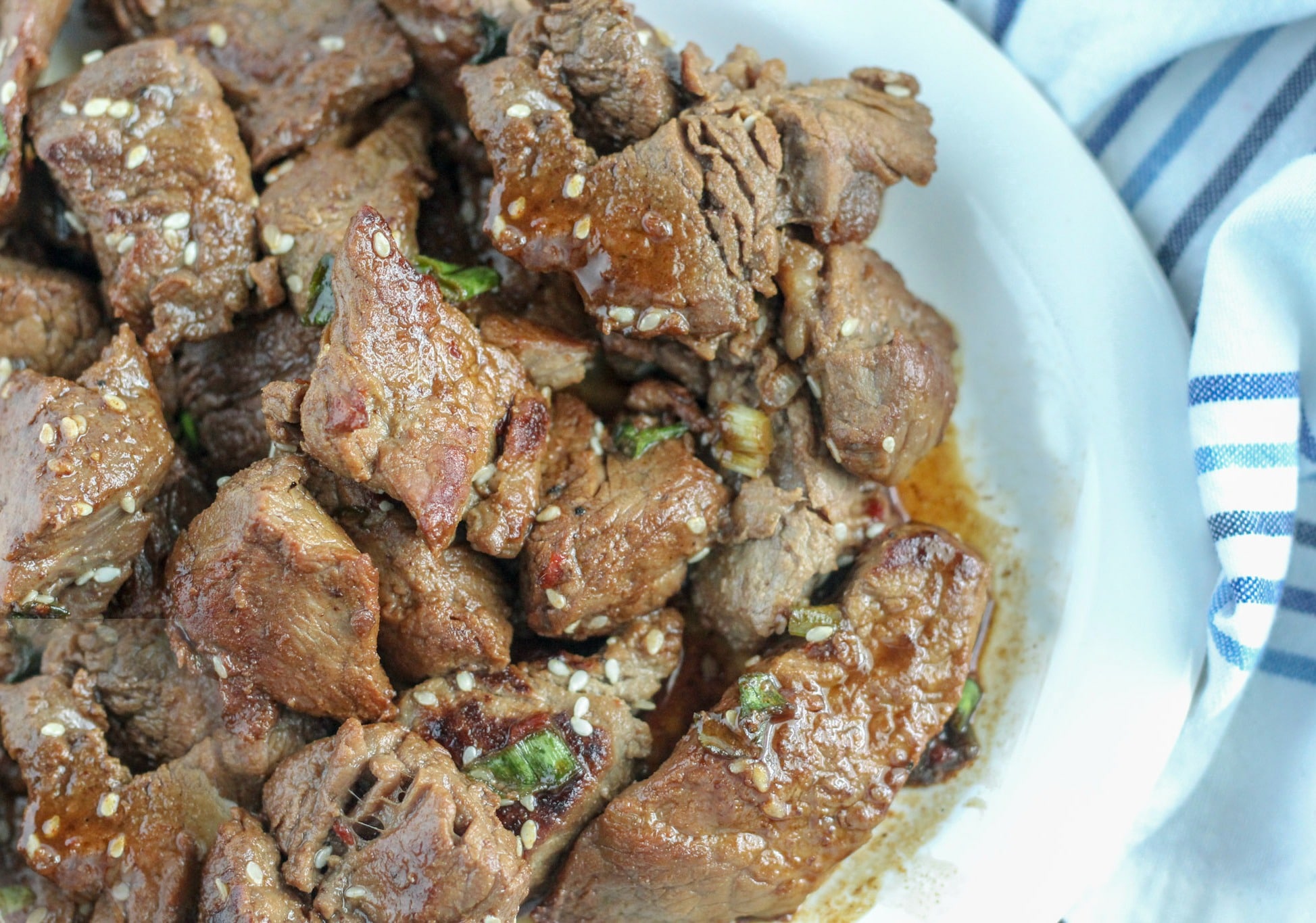 Teriyaki Steak Bites garnished with scallions and sesame seeds on white platter with blue and white napkin 