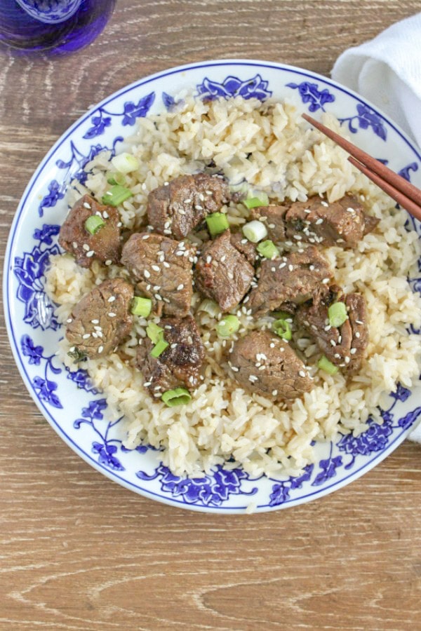 Teriyaki Steak Bites on top of rice with with sesame seeds and scallions on blue and white plate with chop sticks. 