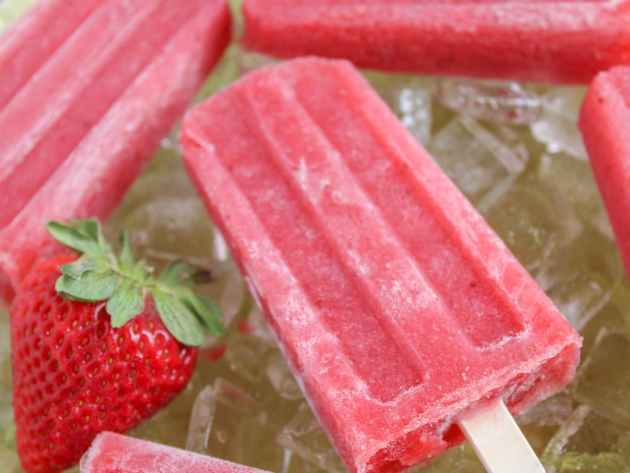 Strawberry Daiquiri Popsicle on wooden stick laying on a bed of ice next to ripe strawberry 