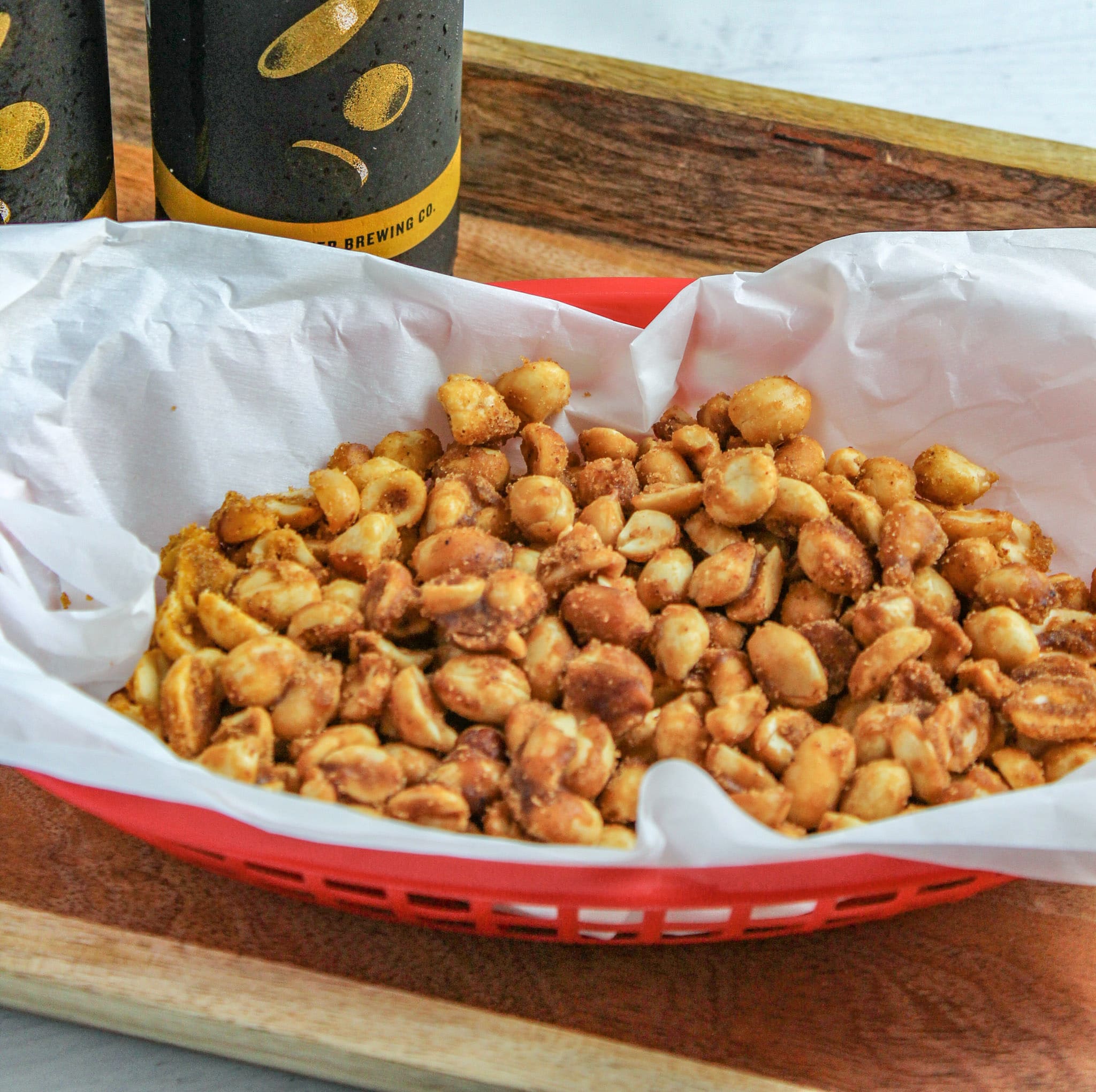 Slow Cooker Sweet and Spicy peanuts in a red plastic basket with white parchment with two bottles of beer on a woodent tray 