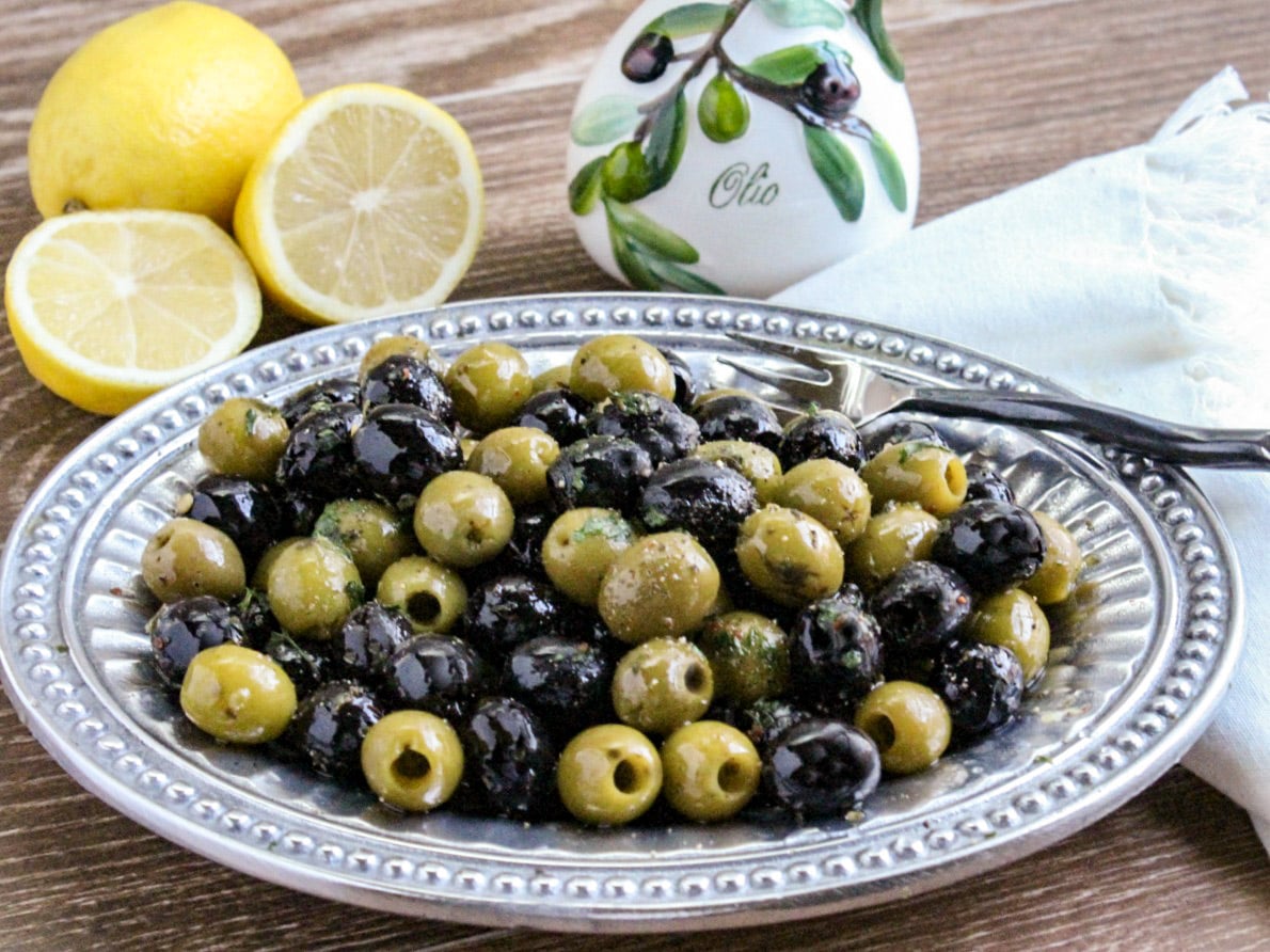 Mixture of green and black pitted olives on a pewter platter with sliced lemons and bottle of olive oil in background 