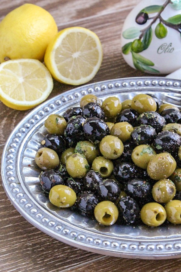 Close up of Garlic and Herb Olive Appetizer on platter with lemons and olive oil bottle in background 