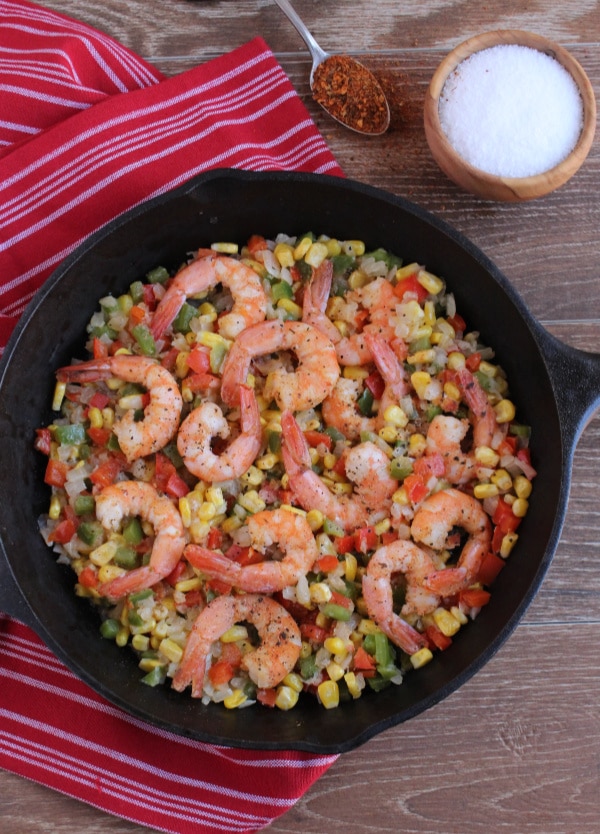 cajun seasoned shrimp with chopped onions and bell peppers and corn in a cast iron skillet on top of a red and white striped towel on wood background 