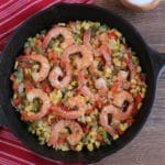 cajun seasoned shrimp with chopped onions and bell peppers and corn in a cast iron skillet on top of a red and white striped towel on wood background