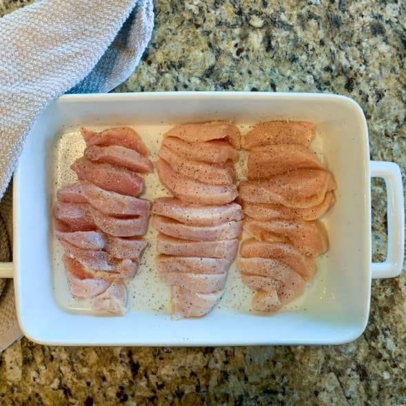 three raw chicken breasts seasoned with salt and black pepper sliced horizontally inside a white baking dish with tan kitchen towel on left on tan granite countertops 