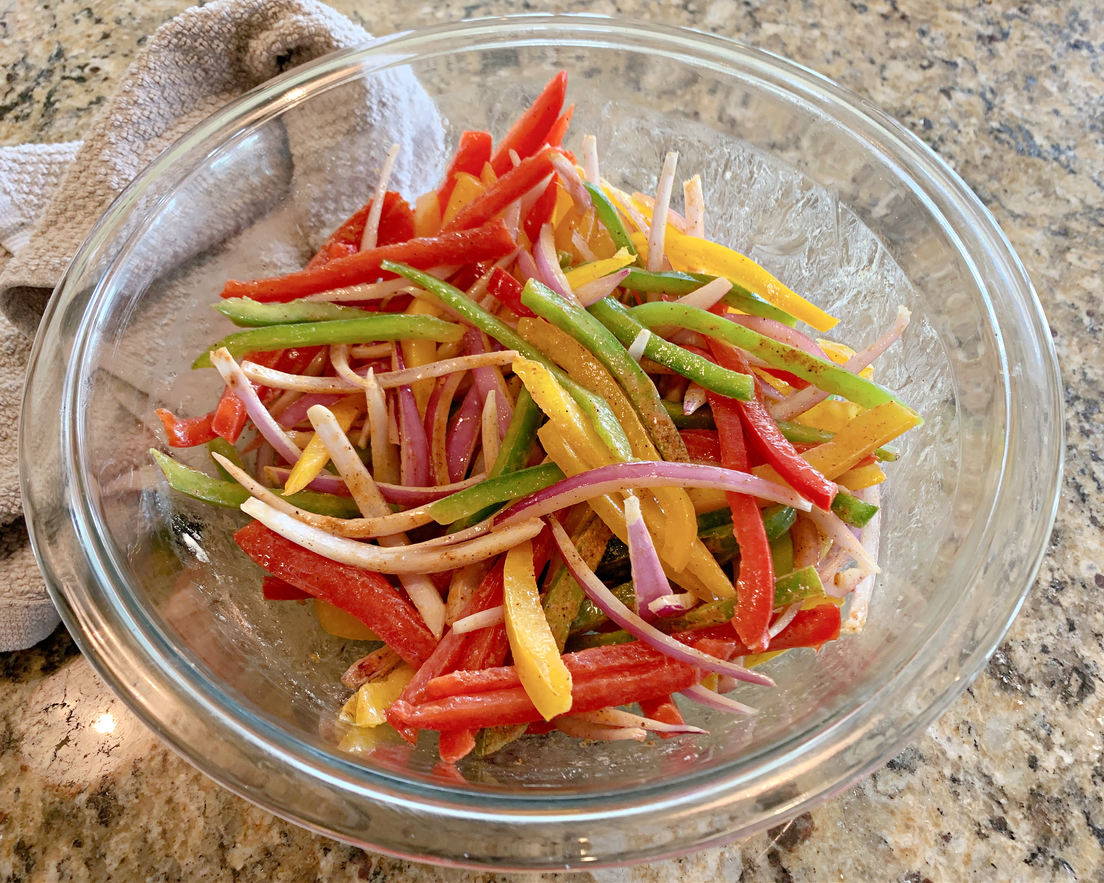 Sliced onions and bell peppers tossed in olive oil and fajita seasoning in a glass bowl. 