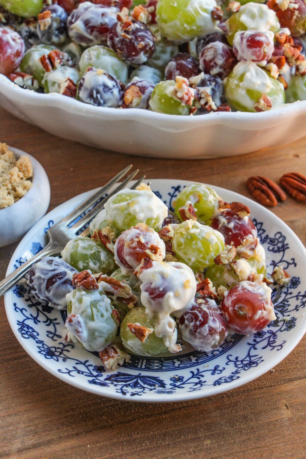 A single serving of Grape Salad on a blue and white plate sits in front of a white serving bowl of the same salad.