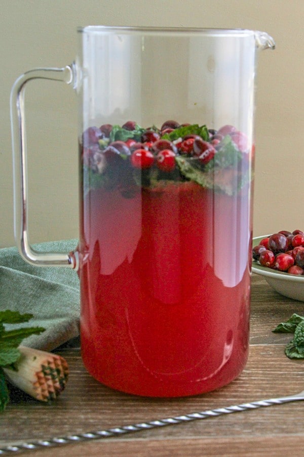 Cranberry Mojitos in a large glass pitcher with mint and cranberries shown with bar spoon, muddlers and fresh cranberries in a bowl