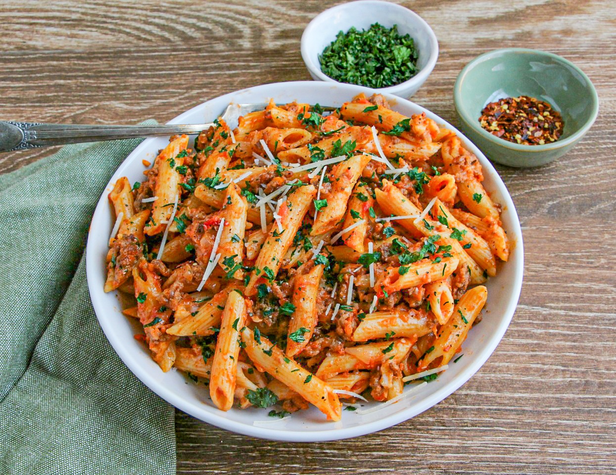 Mostaccioli Pasta garnished with cracked pepper and parsley in a white bowl with green napkin next to it.