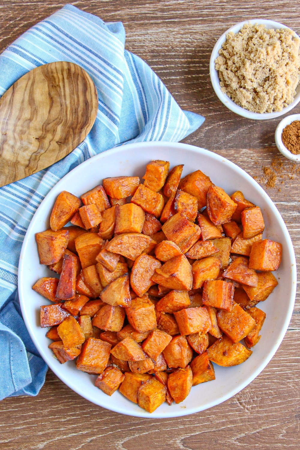 Cinnamon and Brown Sugar Sweet Potatoes in a white bowl on wooden cutting board with blue dish towel and spoon 