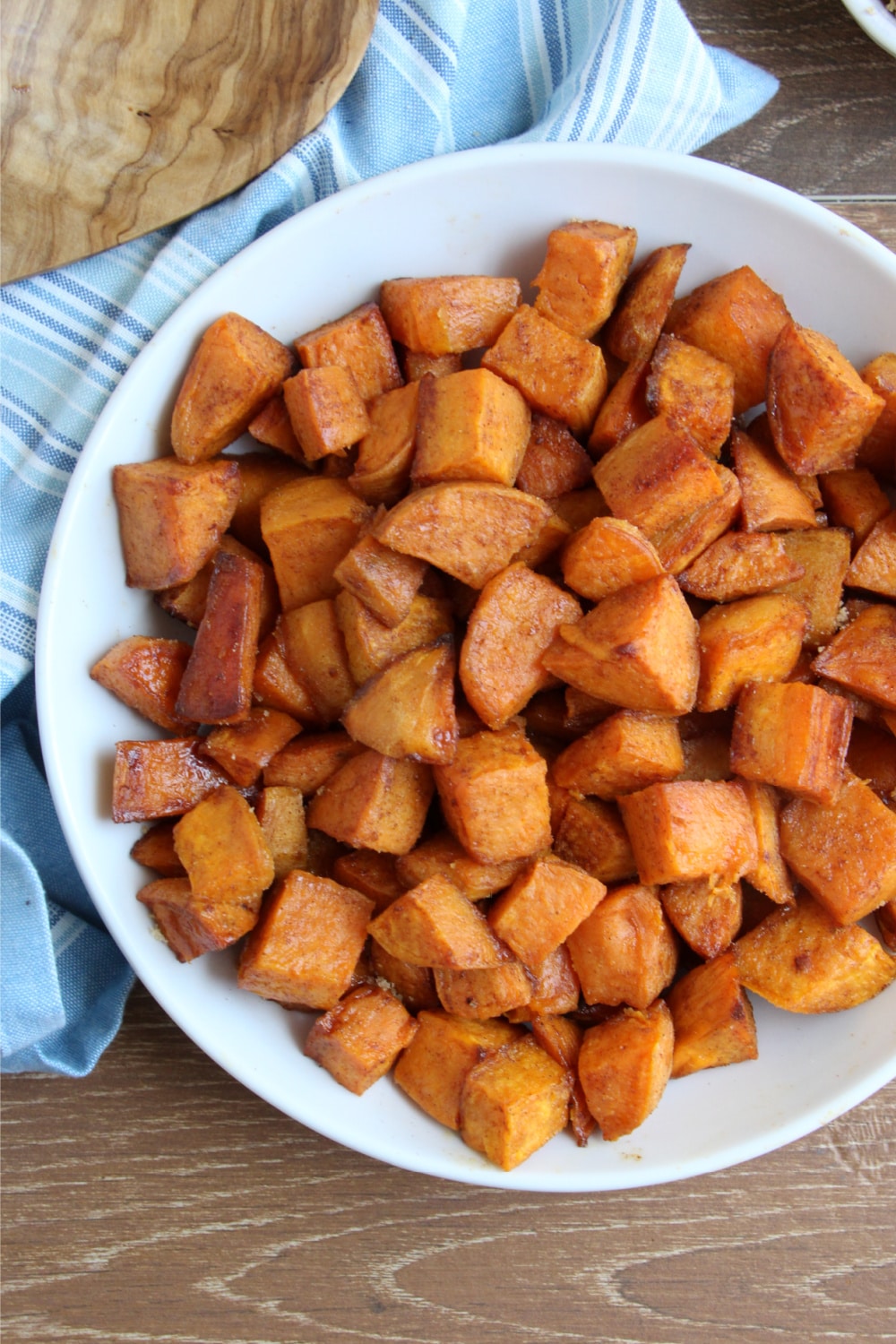 Cinnamon and Brown Sugar Sweet Potatoes in a white bowl on wooden cutting board with blue dish towel and spoon 