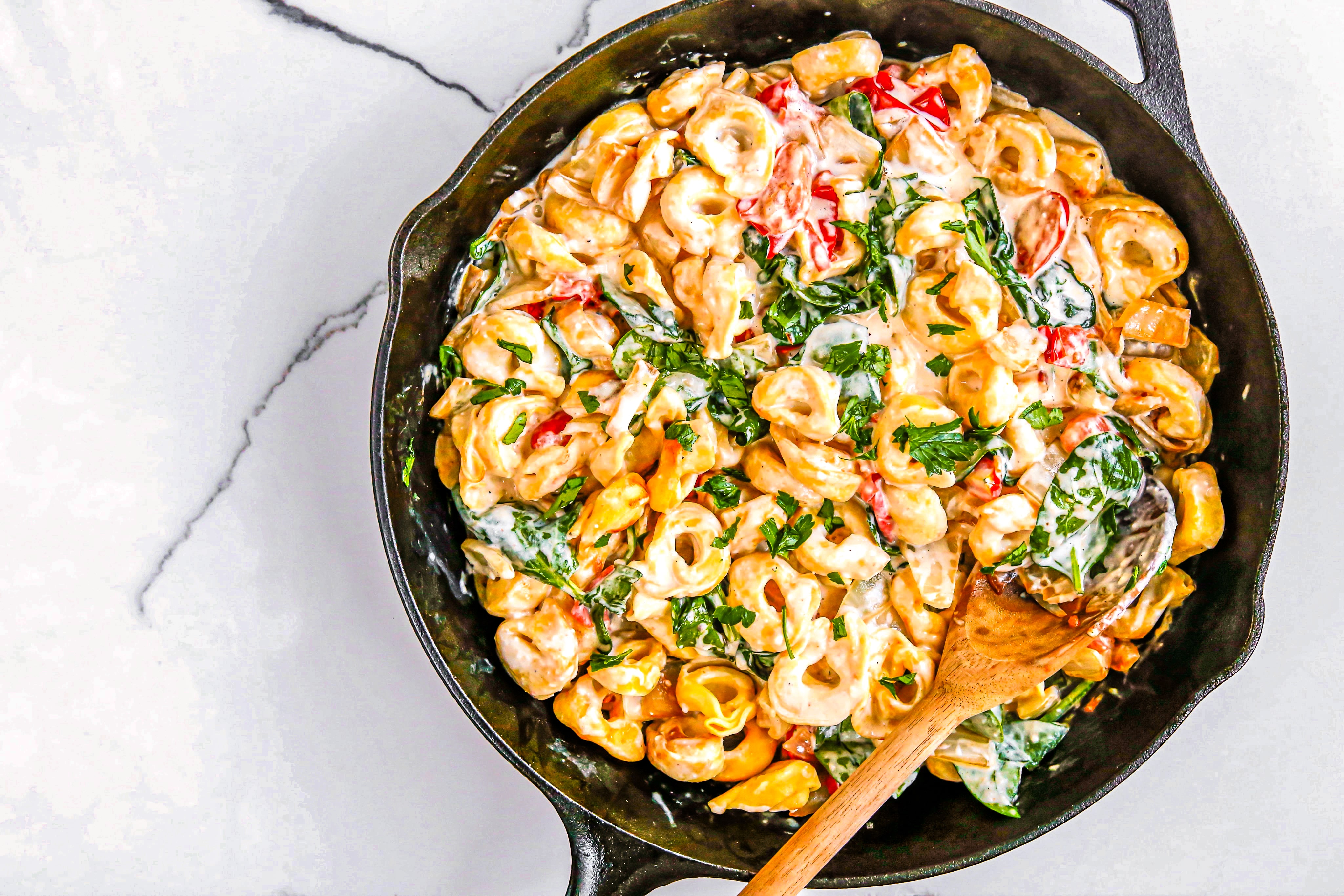 A white bowl of Creamy Spinach Alfredo Tortellini in a cast iron skillet with wooden spoon.