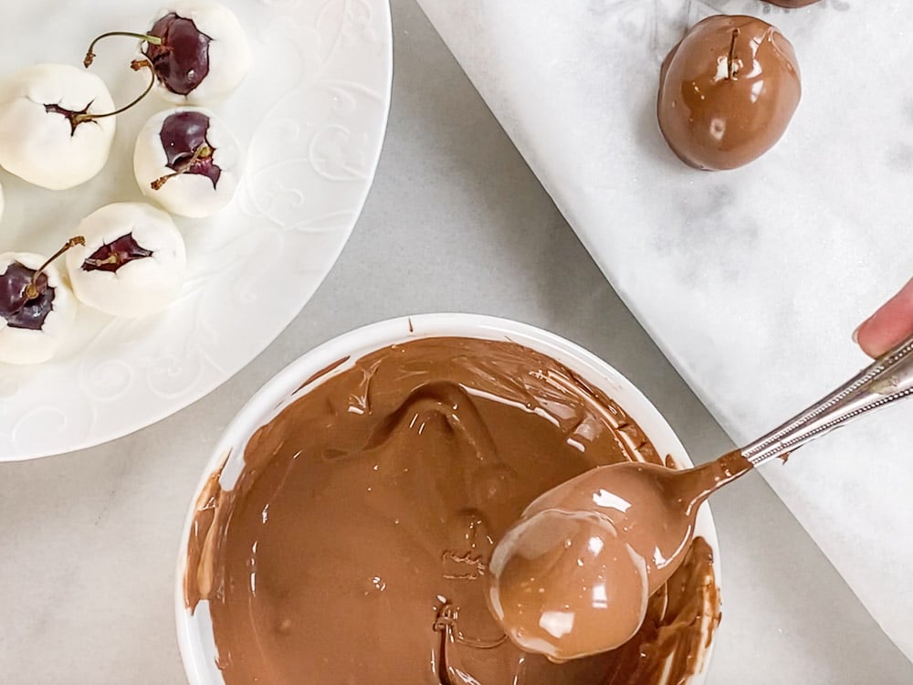 Cherry warped in dough and covered in melted chocolate on a spoon over bowl of melted chocolate 