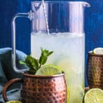 A pitcher of Moscow Mules behind a copper mug garnished with lime and mint.