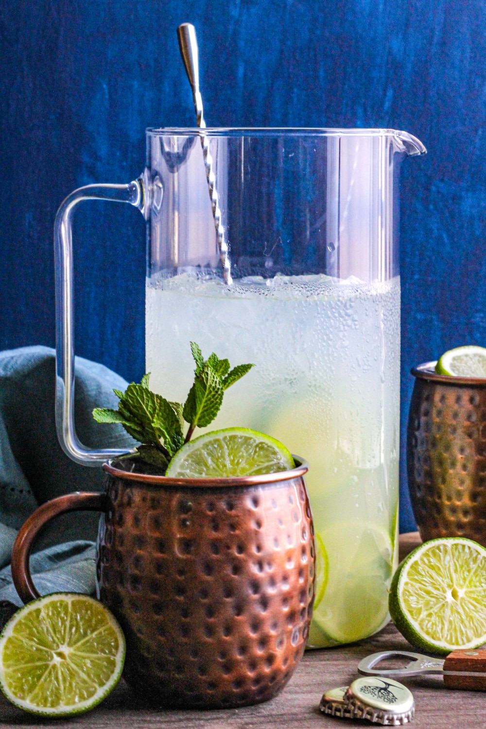 A pitcher of Moscow Mules behind a copper mug garnished with lime and mint. 
