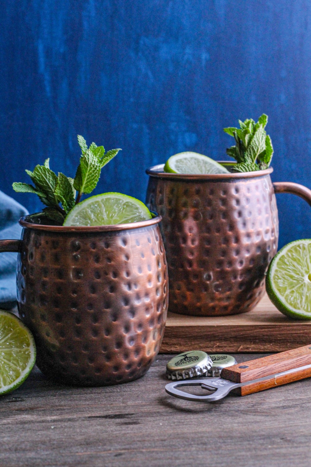 Two Moscow Mules in copper mugs with limes and a bottle opener in the foreground.