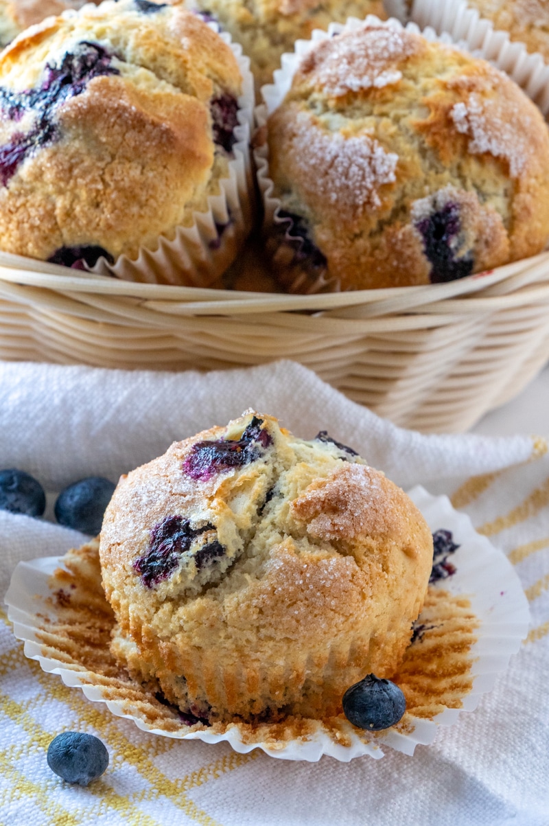 A single blueberry muffin sits in front of a basket of muffins.