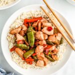 Chicken stir-fry over rice in a white bowl with chop sticks resting on edge of the bowl.
