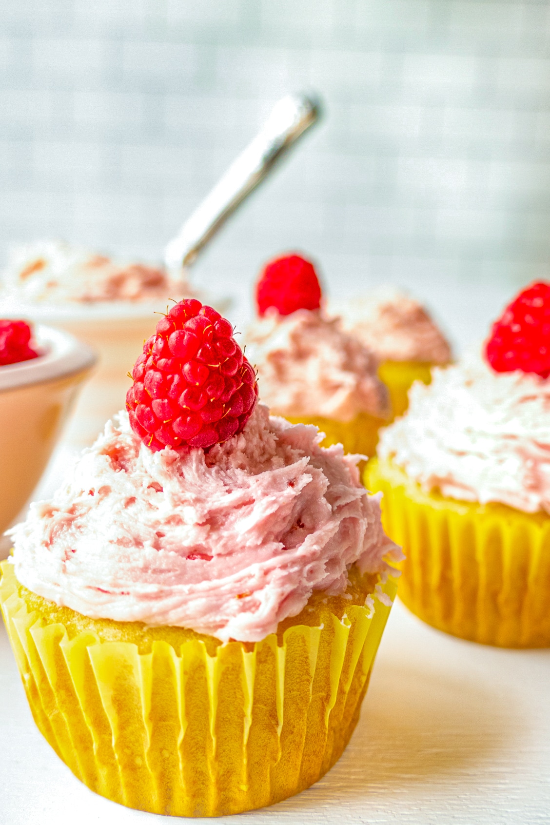 Frosted cupcakes topped with a raspberry sitting in front of a bowl of frosting. 