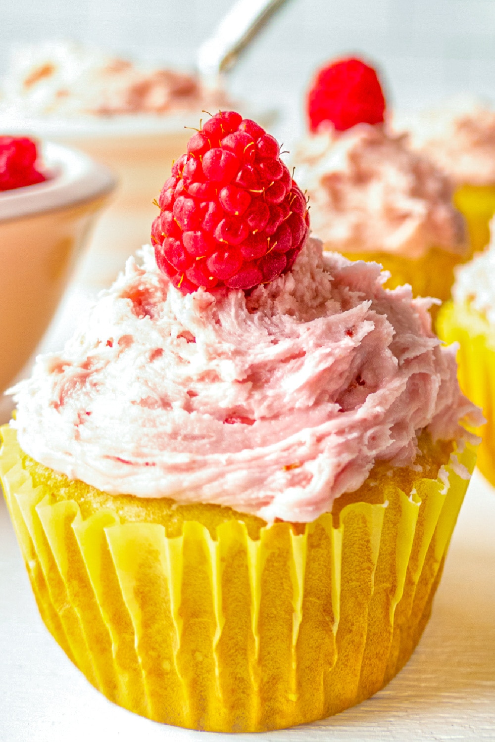 Frosted cupcakes topped with a raspberry sitting in front of a bowl of frosting.
