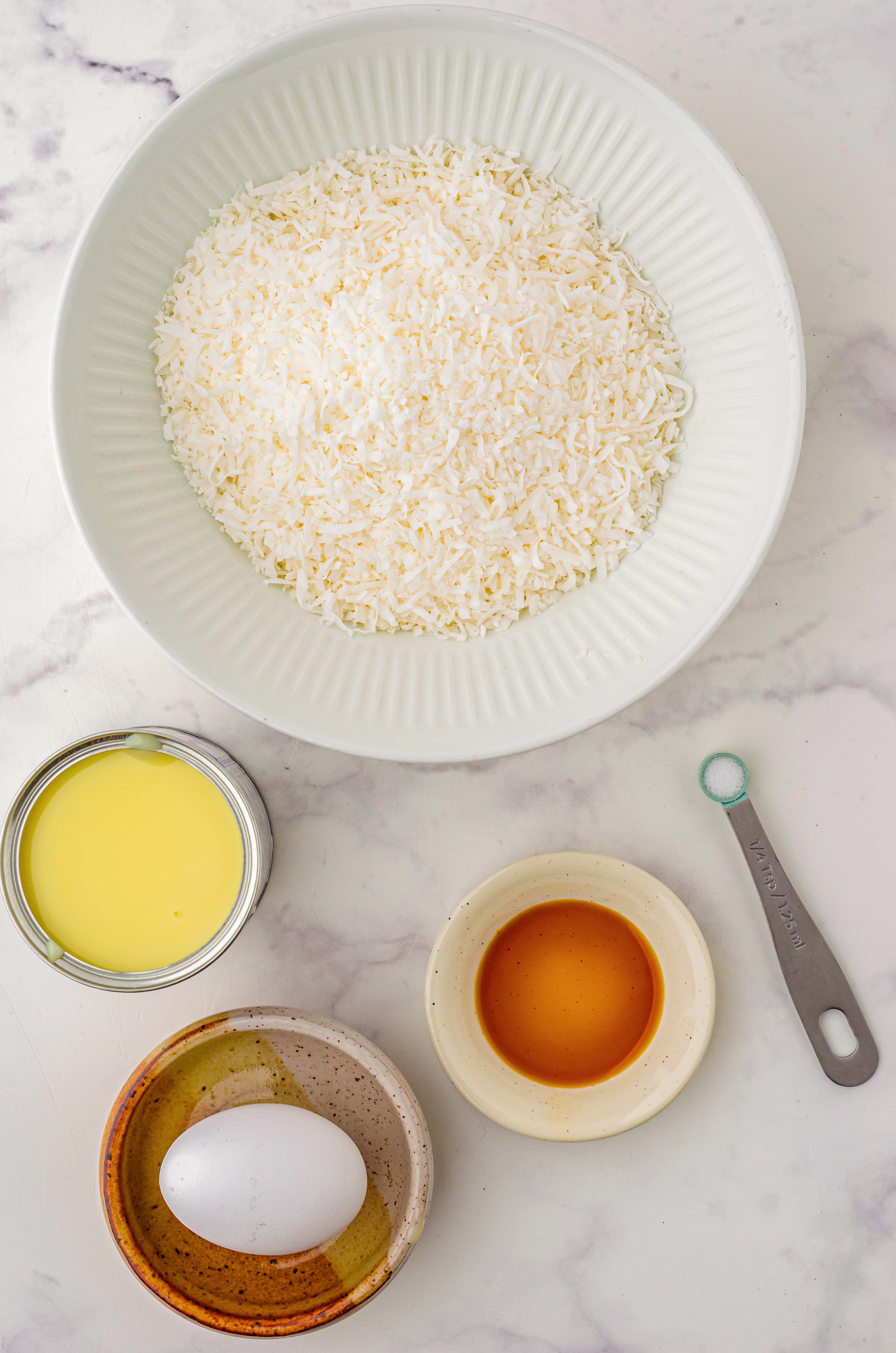  Measured ingredients needed to make Coconut Macaroons laid out in bowls on a marble countertop.