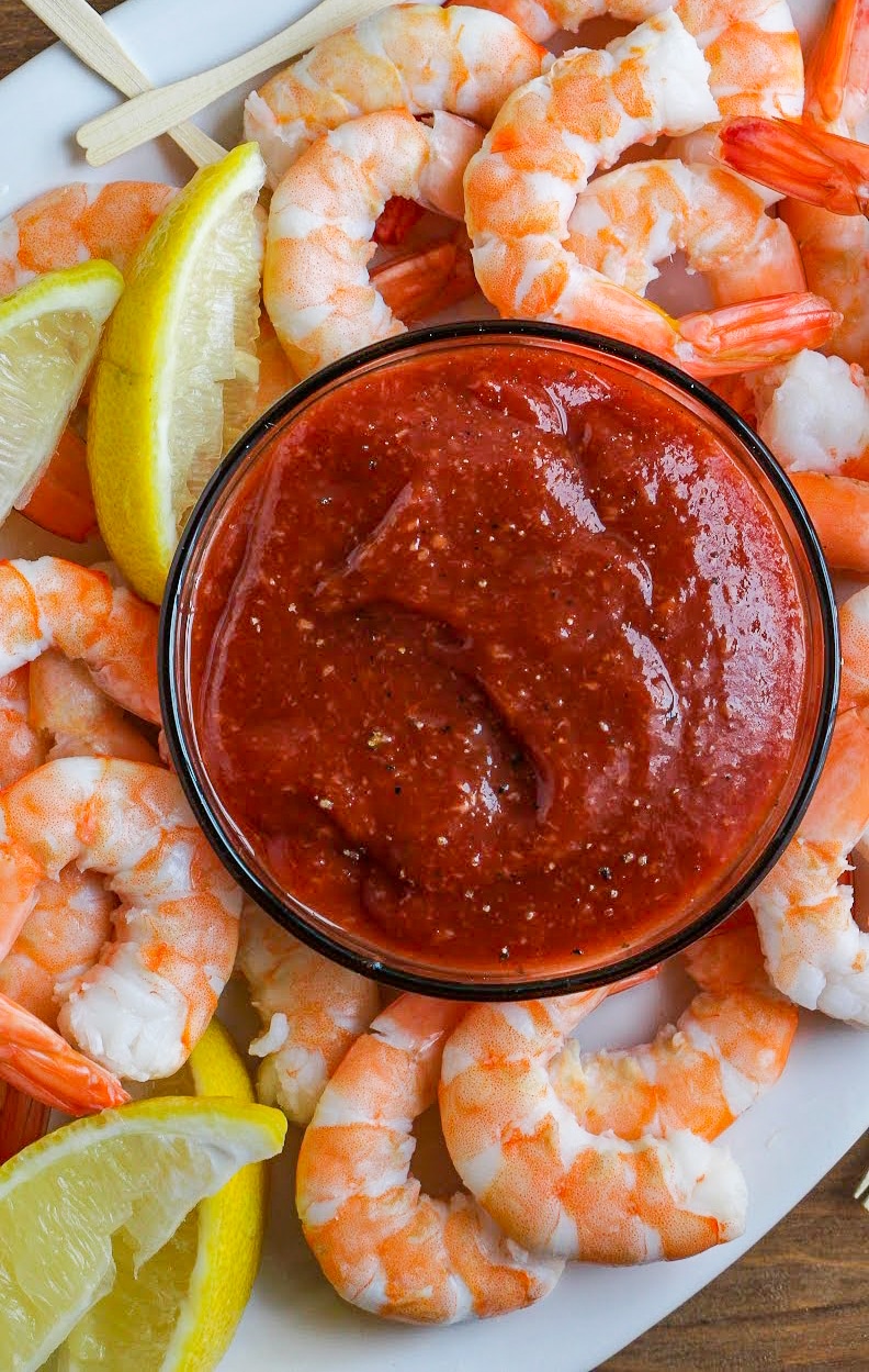 Homemade cocktail sauce in a clear glass bowl surrounded by shrimp and lemon wedges. 
