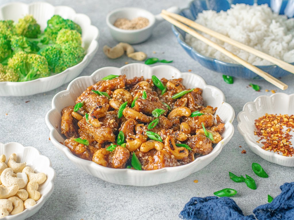 A white bowl of Cashew Chicken sits next to a bowl of steamed broccoli and a bowl of rice.