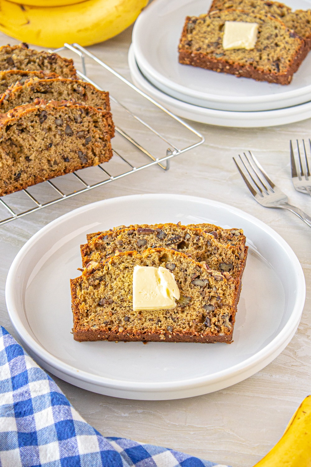 Two slices of banana bread with a pat of butter on a small white plate next to forks on a white counter. 
