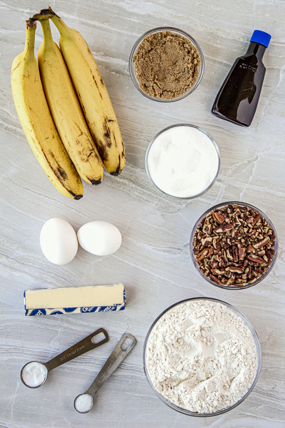 Measured ingredients needed to make Banana Nut Bread in clear glass bowls and aluminum measuring spoons on a white counter top. 