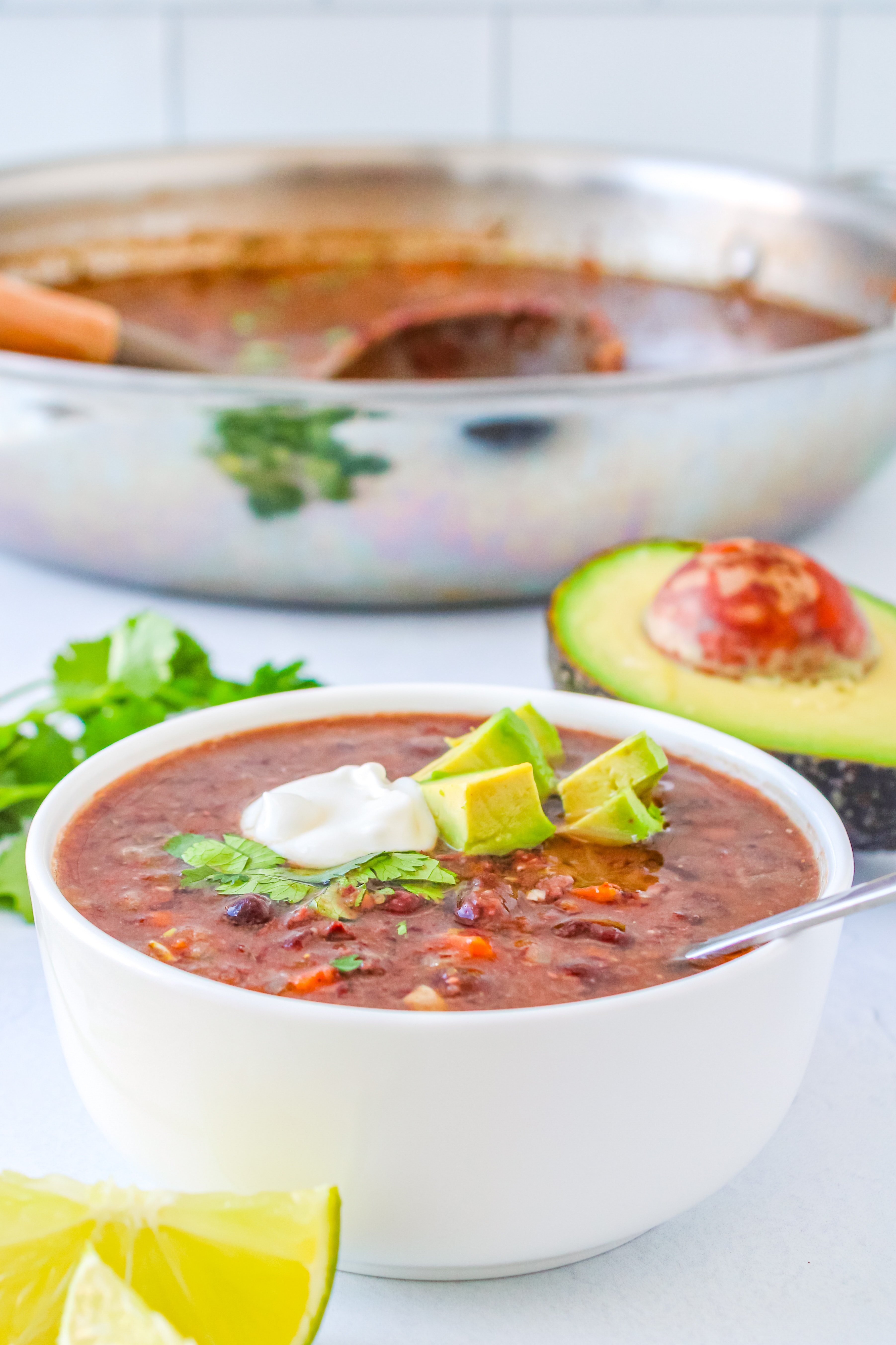 A white bowl filled with black bean soup sits in front of a stainless Steele pot of soup with a ladle inside. 