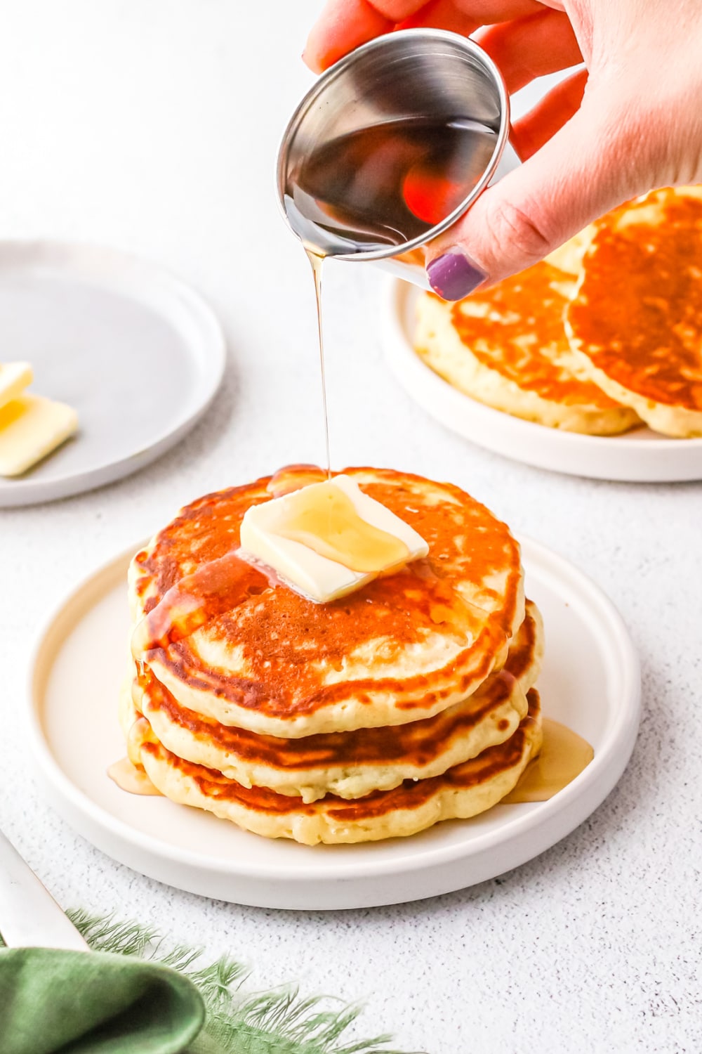 A small stack of pancakes topped with butter and syrup being poured over the top by a woman's hand.
