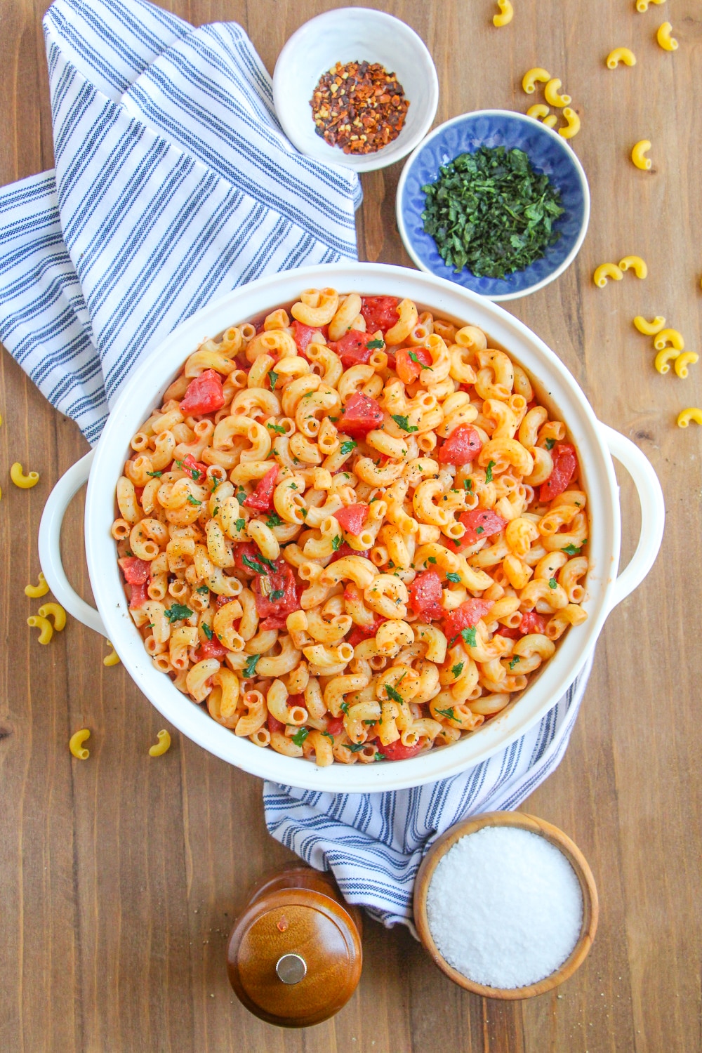 Macaroni and tomatoes in a white serving bowl with handles on a blue and white dish towel. 