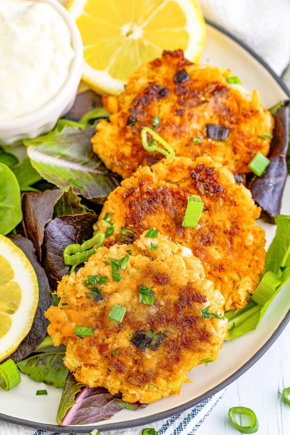 Three salmon patties lay on a bed of greens next to lemon slices and a cup of tater sauce.
