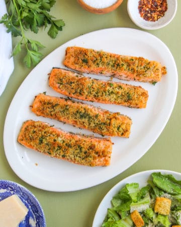 Four Garlic Parmesan Salmon fillets on a white platter sitting on a green background.
