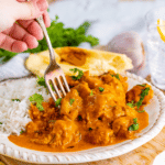 A woman's hand holding a fork stabs a piece of butter chicken garnished with cilantro.