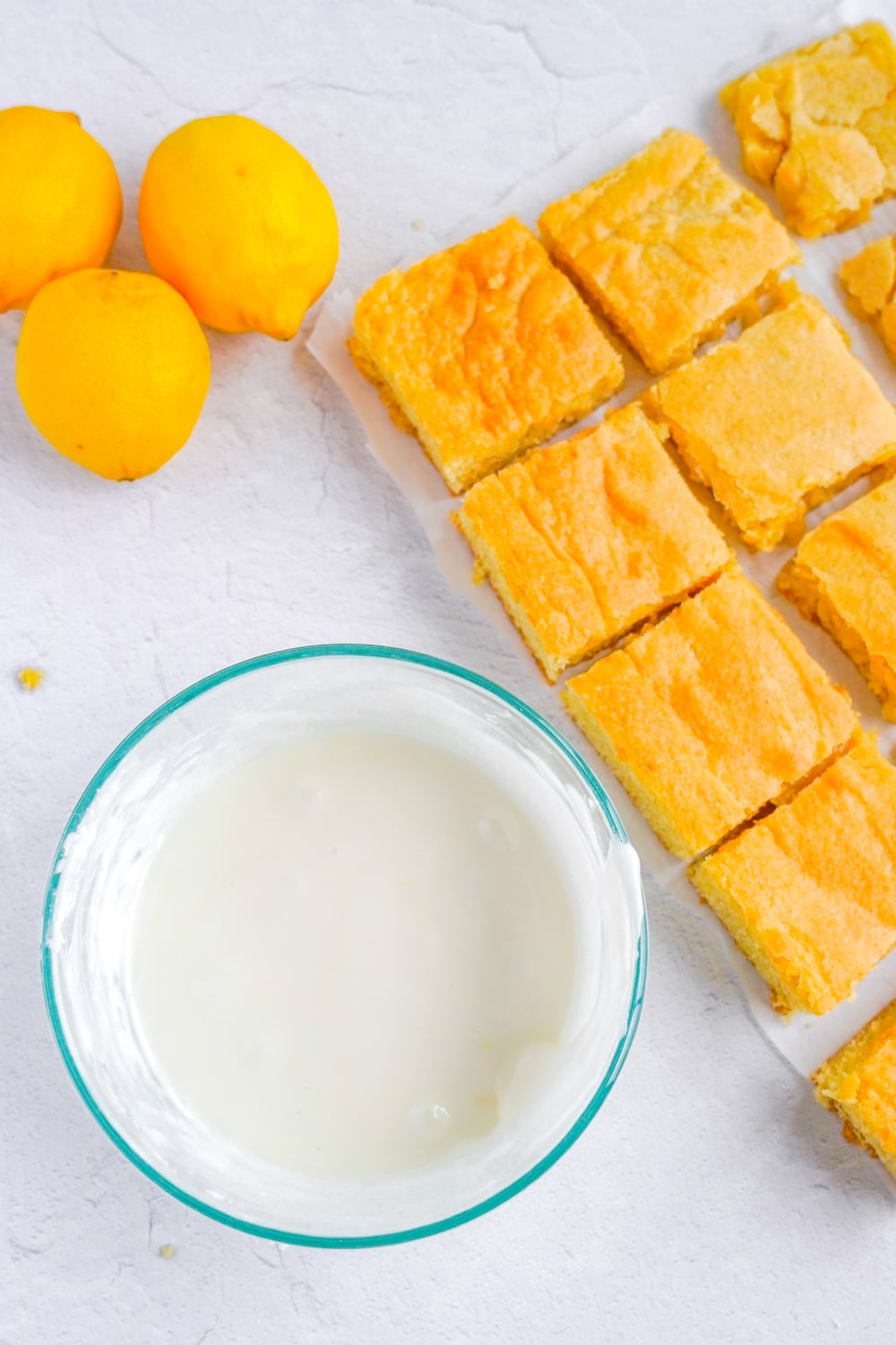 A clear glass bowl filled with lemon sugar glaze sits next to sliced unglazed cookie bars and three lemons.
