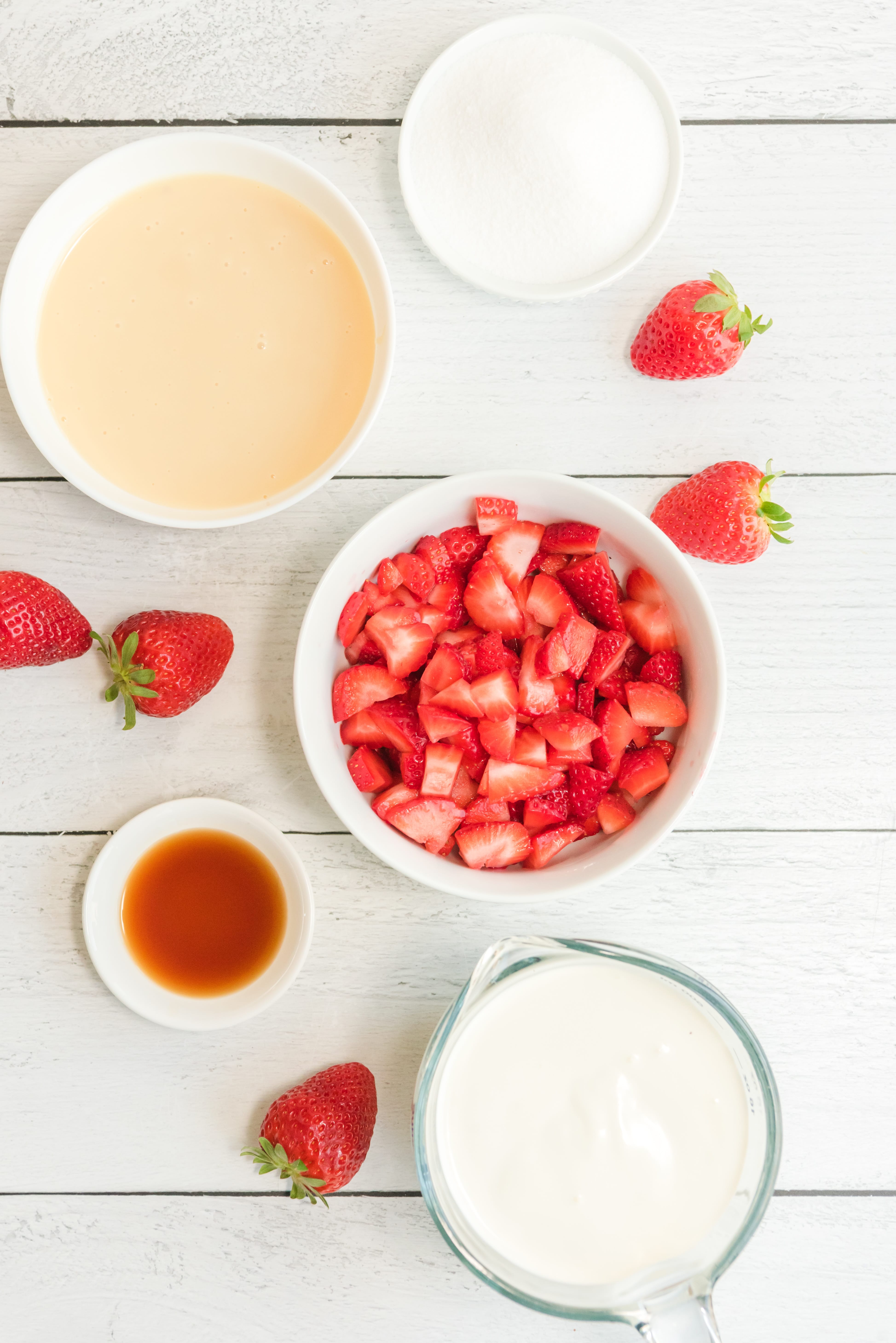 Ingredients needed to make No Churn Strawberry Ice Cream presented in white bowls on a white shiplap background accented with fresh berries. 