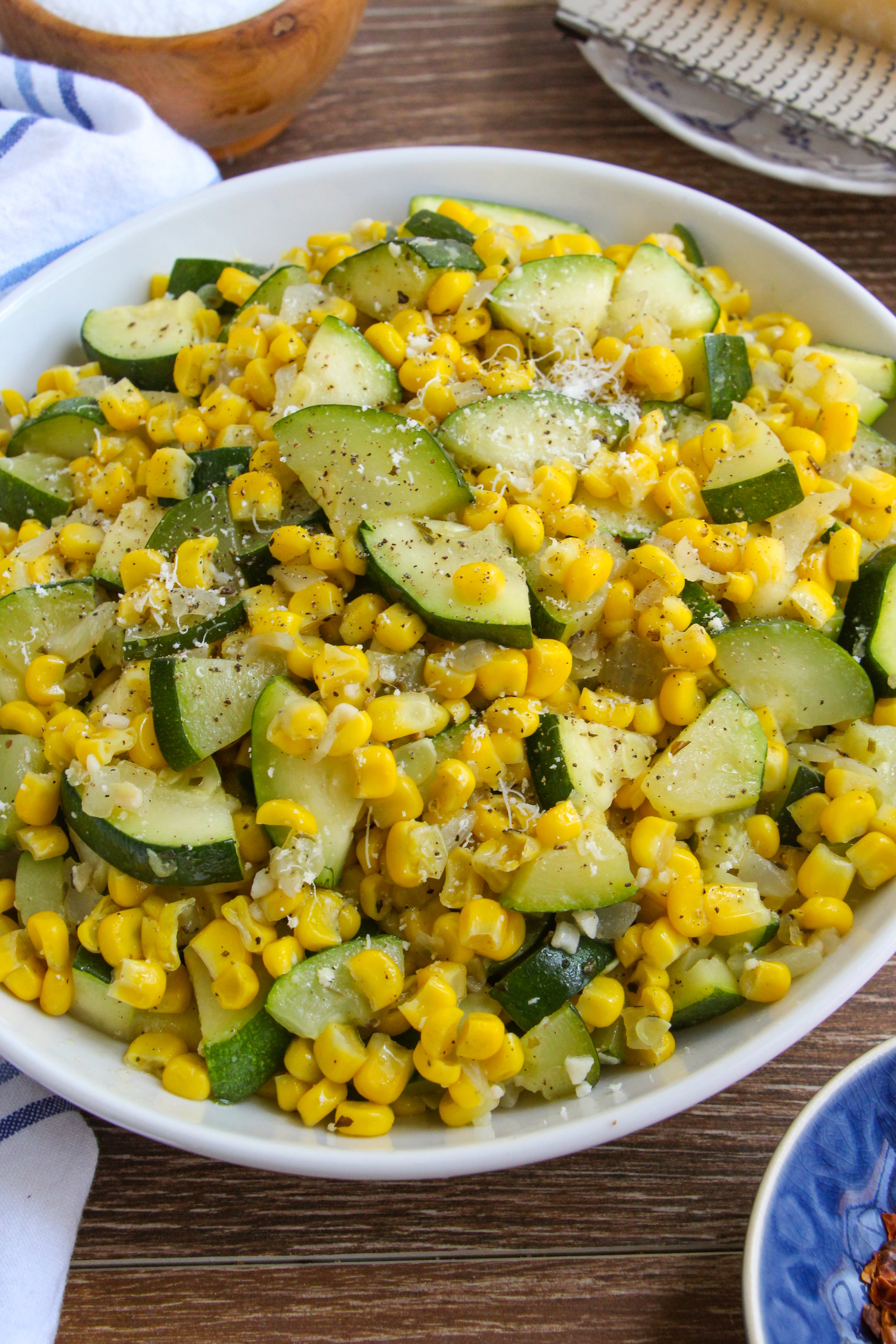 Zucchini and corn sauté in a white bowl on a wood tabletop next to a block of parmesan cheese and grater. 