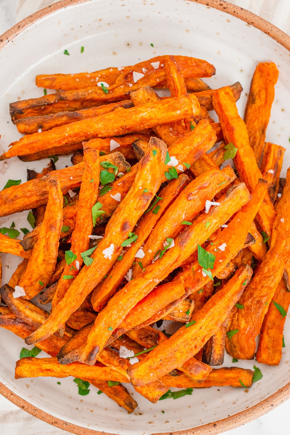 A white plate piled high with Baked Sweet Potato Fries granshied with flaky salt and parsley.