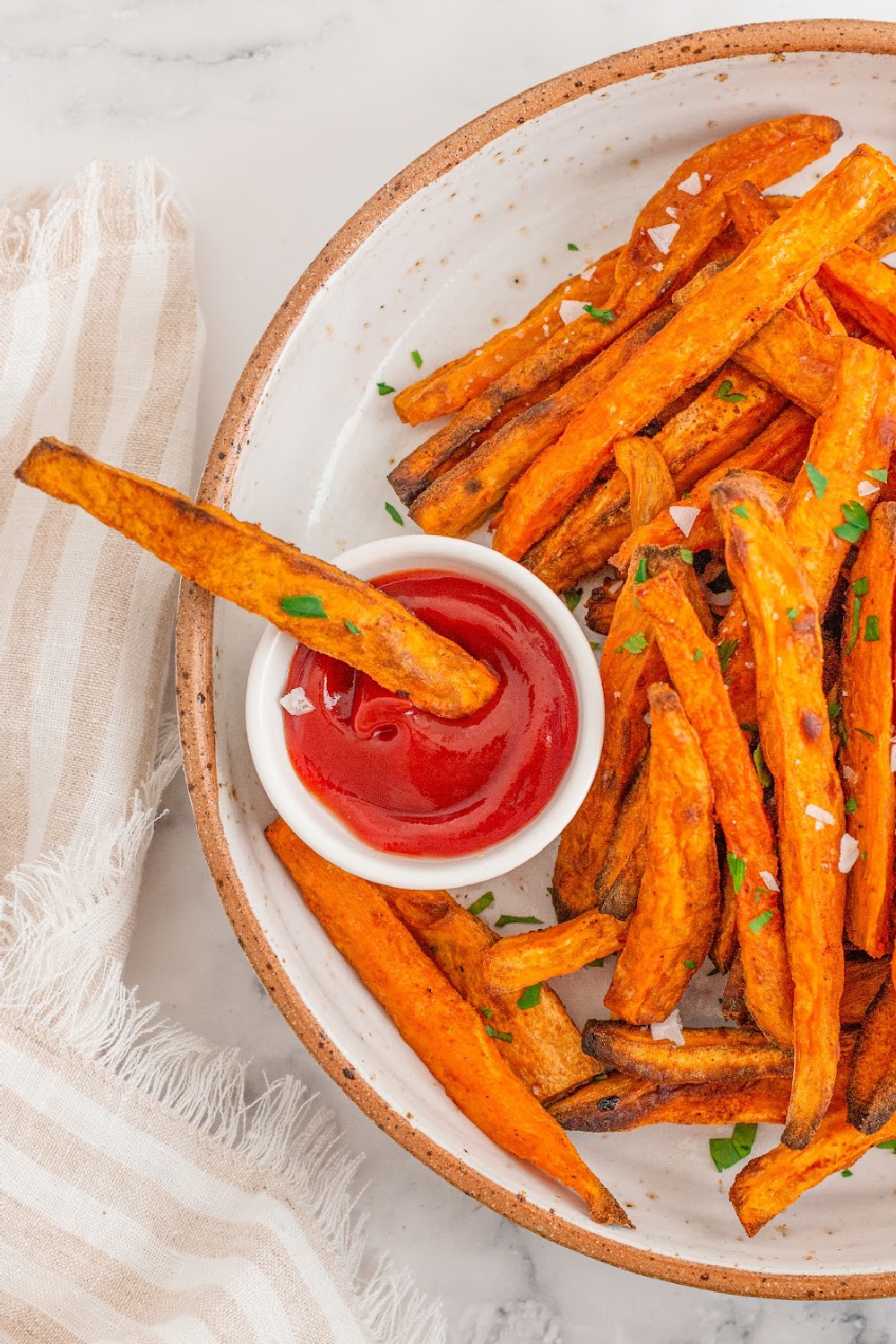 Sweet potato fries on a white plate sit next to a white cup of ketchup with a single fry dipped inside it. 