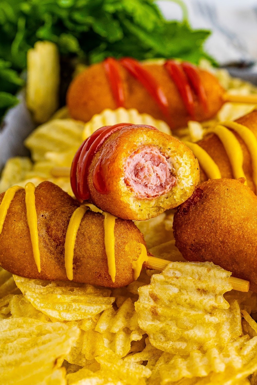 Mini Homemade Corn Dogs some drizzled with ketchup or mustard lay on a bed of potato chips. One corn dog is missing a bite showing both the cornmeal coating and the hotdog inside.