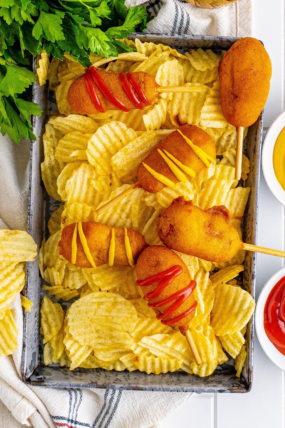 Six corn dogs in a metal tray sitting on a pile of chips next to small white bowls filled with ketchup and mustard.