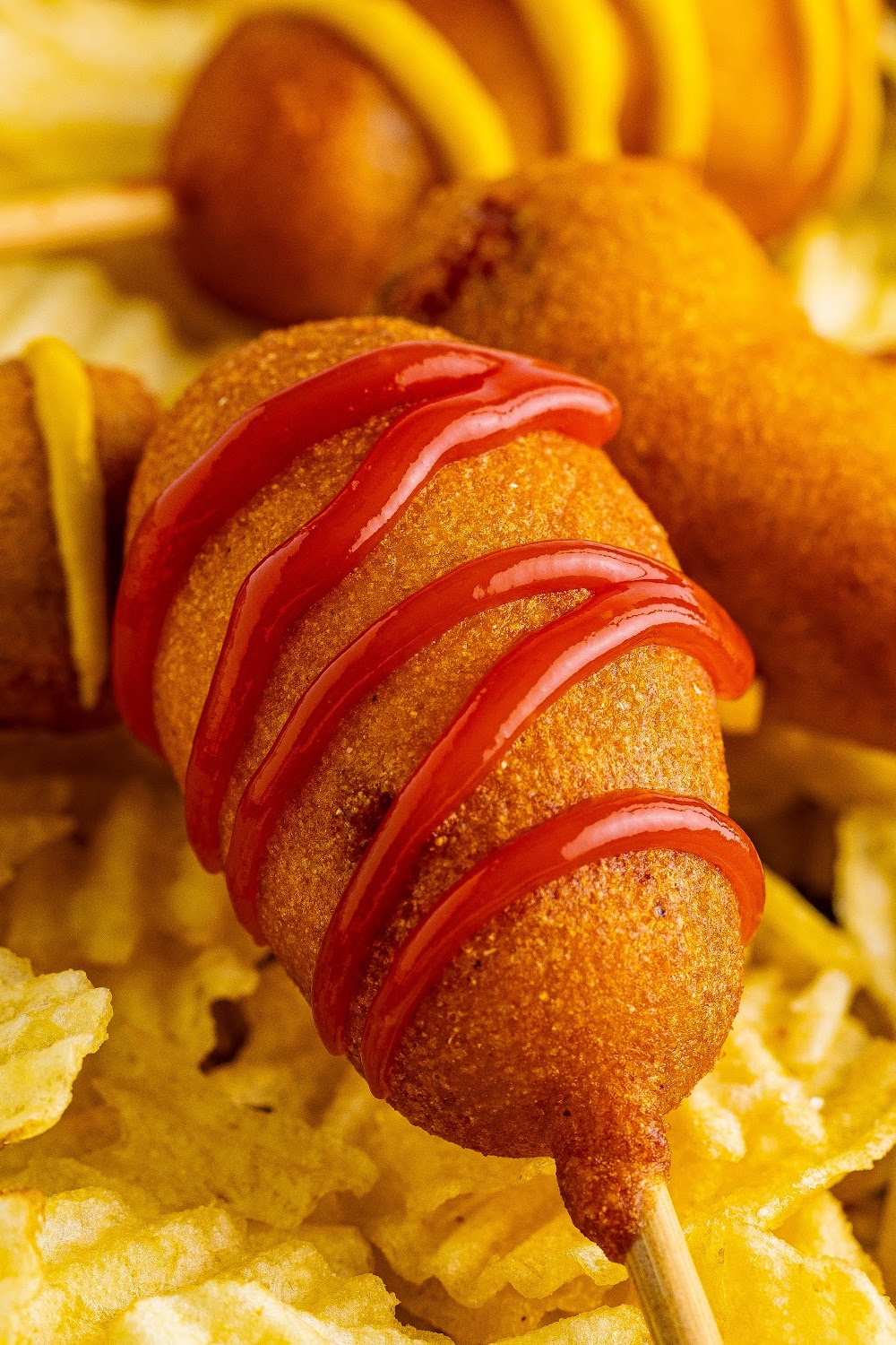 A corn dog drizzled in ketchup with a bamboo skewer sits a top a bed of potato chips.